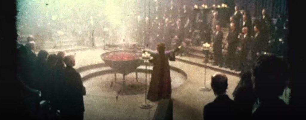 A grainy image of a religious ritual is seen. A crowd of people dressed in all black encircle Christabella. In front of Christabella is a blackened child being burned alive over hot coals.