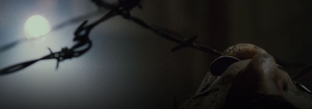 The face of a corpse is seen looking up at the ceiling with a key sticking out of its mouth. There is barbed wire holding it in place.