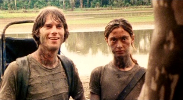 A white man covered in mud is seen looking at what appears to be a body almost completely offscreen and grinning while standing next to a while woman frowning at the same thing. There is water and the edge of a jungle in the background.