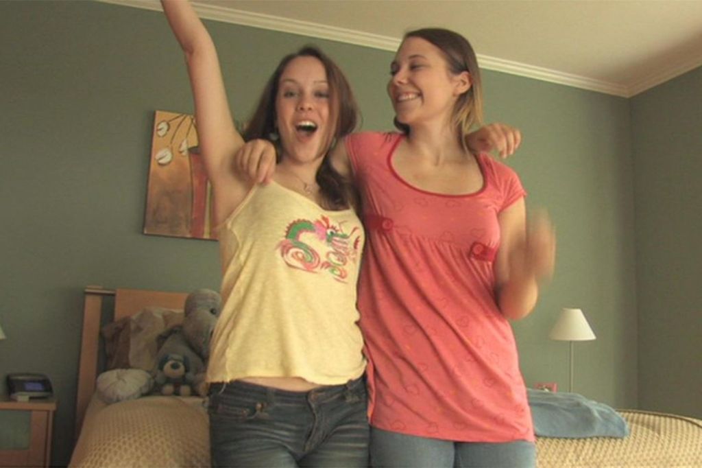 Two white girls are standing with their arms around each other smiling in front of a bed.