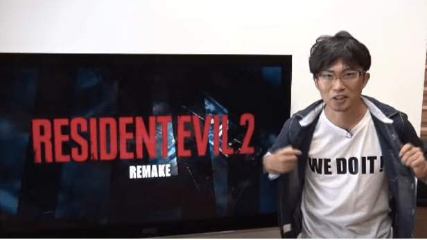 Image of a Japanese man standing in front of a TV monitor that says "Resident Evil 2 Remake". He is holding his jacket open to reveal a white shirt with black lettering that says "We do it!"